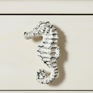 May include: A white seahorse-shaped cabinet knob with black distressed detailing. The knob is mounted on a white cabinet. This decorative hardware adds a coastal or nautical theme to furniture.
