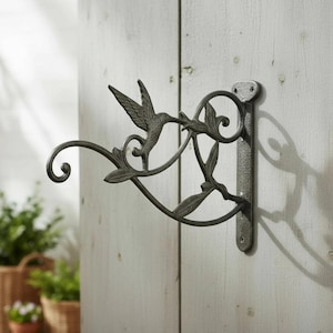 May include: A decorative, cast iron hummingbird bracket in a dark gray finish. The bracket features a hummingbird design with leaves and a curved arm for hanging items. It is mounted on a wooden surface.