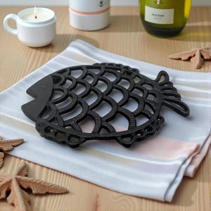 May include: A black cast iron trivet in the shape of a fish, resting on a striped cloth. The trivet features a detailed scale pattern and fins. A lit candle and other decorative items are in the background.
