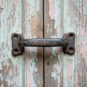 May include: A weathered, dark metal handle with the number "1388" embossed on it, mounted on a wooden surface with peeling paint in shades of pink, blue, and brown. The handle has a rustic, vintage appearance.