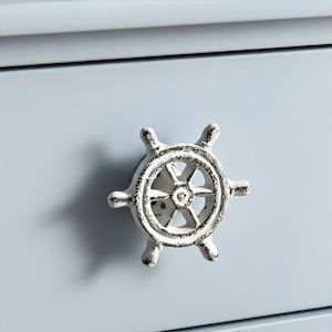 May include: Close-up of a white, ship's wheel-shaped cabinet knob with a distressed finish. The knob is mounted on a light blue cabinet door. Nautical-themed hardware.
