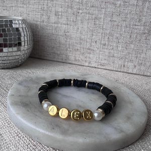 Może przedstawiać: Bransoletka z czarnych koralików z złotymi koralikami literowymi, które tworzą napis "ALEX" i białymi akcentami pereł.