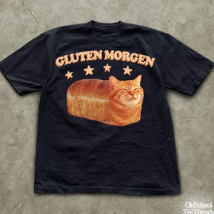 Puede incluir: Camiseta negra con la frase "GLUTEN MORGEN" en naranja sobre un pan con forma de gato naranja sonriente. El diseño incluye cinco estrellas naranjas. La camiseta es de OldSchool TeeThreads.