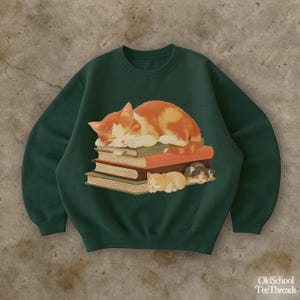 Pode incluir: Um moletom verde escuro com um gráfico de um gato laranja e branco dormindo em cima de uma pilha de livros. Dois gatos menores dormindo embaixo. O texto "OldSchool TeeThreads" está no canto inferior direito.