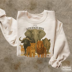 Pode incluir: Camisola creme com um gráfico de vários animais de safari, incluindo um elefante, um leão e um rinoceronte. A palavra "SAFARI" está acima da palavra "ADVENTURES" em negrito e em preto. A camisola tem mangas compridas e gola redonda.
