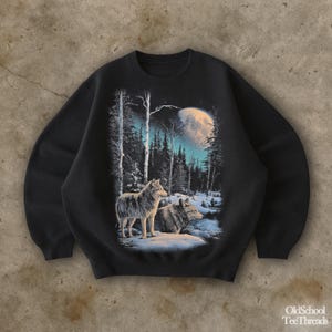 May include: Black sweatshirt with a graphic of two wolves in a snowy forest under a full moon. The design includes tall trees and a wintery landscape. The sweatshirt has a classic crew neck.