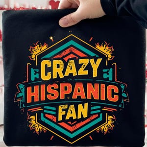 Puede incluir: Sudadera negra con un diseño gráfico colorido. El diseño presenta el texto "CRAZY HISPANIC FAN" en naranja, amarillo y turquesa, dentro de una forma geométrica con detalles de llamas. La sudadera es sostenida por una mano.