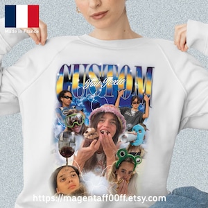 May include: White sweatshirt with a graphic design featuring the word "CUSTOM" in a stylized font, with a collage of images and the text "You Texie". The sweatshirt has a crew neck and long sleeves. The French flag is visible in the upper left corner.