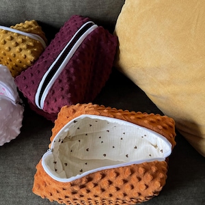 Puede incluir: Una bolsa abierta de tela minky naranja con cremallera blanca y un interior blanco estampado con pequeños corazones marrones. Se ven otras bolsas minky en burdeos y dorado.
