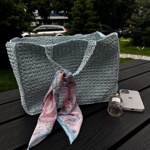 May include: A light blue crocheted tote bag with matching handles. A pink floral scarf is tied to the handle. A bottle of perfume and a white smartphone are next to the bag on a dark wooden surface.