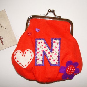 Puede incluir: Una cartera de monedas de color naranja brillante con un corazón blanco, una flor morada y una letra N grande con un patrón floral. La cartera tiene un cierre plateado y está hecha a mano.