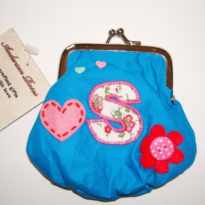 Puede incluir: Una cartera de monedas azul brillante con una aplicación de corazón, flor y letra S. La cartera tiene un marco de metal plateado y un botón rosa con lunares blancos.