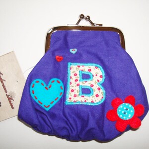 Puede incluir: Monedero morado con una gran letra B blanca con flores rosas, un corazón turquesa y una flor roja con un botón turquesa. El monedero tiene un cierre plateado y una etiqueta que dice "Anderson House, Regalos hechos a mano, Hecho con amor."