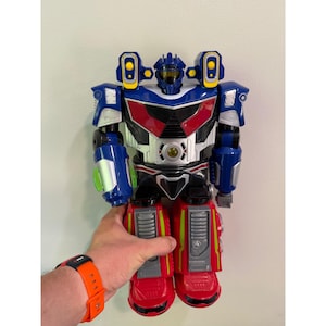 May include: A large, colorful robot toy with a blue head and torso, red accents, and silver details. The robot has large red feet and is held by a hand. The toy has yellow and green highlights.