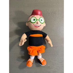 Può includere: Una bambola di peluche raffigurante un personaggio con capelli rossi, occhiali rotondi verdi e un ampio sorriso. Indossa una maglietta nera a maniche corte con una striscia orizzontale arancione, pantaloncini cargo arancioni e scarpe da ginnastica.