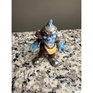 May include: A blue and brown action figure of a fantasy character. The figure has a blue face, arms, and hands, with a brown outfit and silver armor. The figure is standing on a speckled surface.