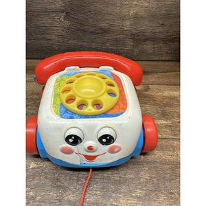 May include: A vintage toy telephone with a red handset and wheels. The white body features a yellow rotary dial and colourful accents. The toy has a smiling face with rosy cheeks and a red pull string.