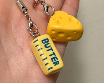 May include: Two food-themed charms: a yellow cheese wedge and a yellow butter stick with blue text that reads "BUTTER". Each charm has a silver clasp and a black cord. The charms are held in a hand.