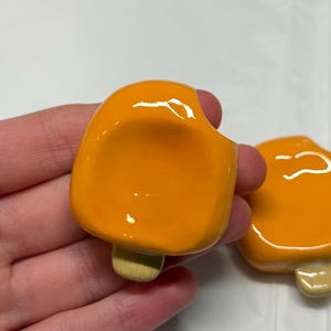 May include: Two orange popsicle-shaped ceramic pins with a bite taken out of one. The pins have a glossy finish and a light brown stick. The pins are likely handmade and suitable for clothing or accessories.