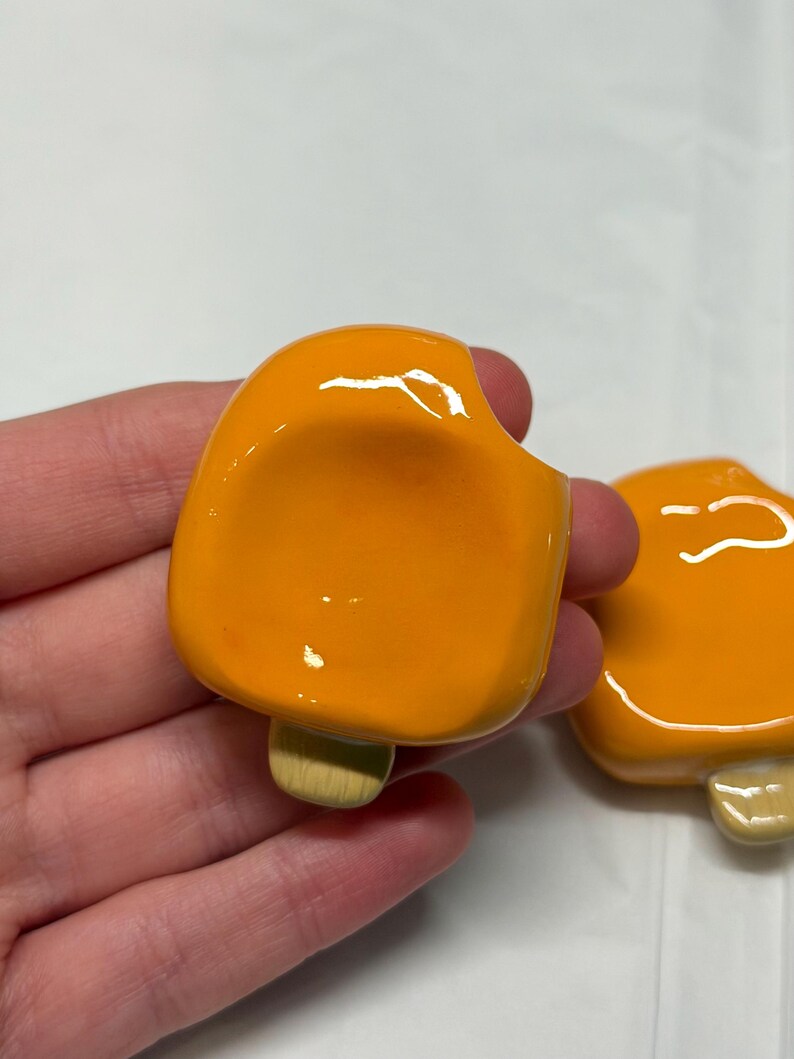 May include: Two orange popsicle-shaped ceramic pins with a bite taken out of one. The pins have a glossy finish and a light brown stick. The pins are likely handmade and suitable for clothing or accessories.