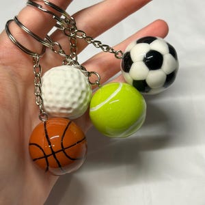 May include: A set of four sports ball keychains featuring a miniature orange basketball with black lines, a white dimpled golf ball, a bright neon yellow tennis ball, and a black and white soccer ball, each attached to a silver metal ring.