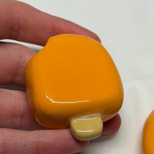 May include: A bright orange popsicle-shaped object with a light yellow stick. The object is smooth and glossy, with rounded edges. The popsicle is held in a hand.