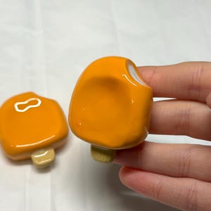 May include: Two orange popsicle-shaped ceramic magnets with white accents and tan stick bases. One magnet is in the foreground, and the other is in the background. The magnets are on a white surface.