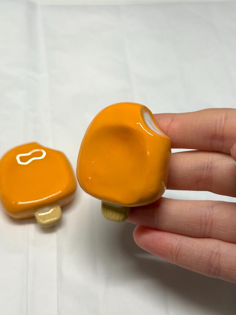 May include: Two orange popsicle-shaped ceramic magnets with white accents and tan stick bases. One magnet is in the foreground, and the other is in the background. The magnets are on a white surface.