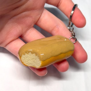 May include: A keychain featuring a miniature, realistic-looking eclair pastry. The eclair has a golden-brown baked dough base, a glossy tan icing glaze on top, and a textured, light-colored interior visible where a bite has been taken out.