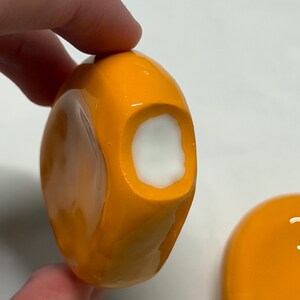 May include: Close-up of an orange, rounded object with a white interior. The object is held by a hand. Another similar orange object with a white smiley face is visible in the background.