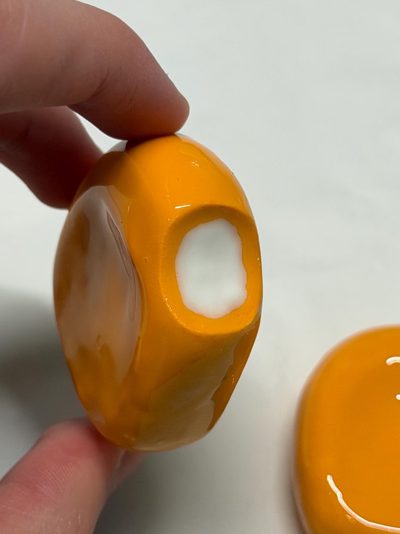 May include: Close-up of an orange, rounded object with a white interior. The object is held by a hand. Another similar orange object with a white smiley face is visible in the background.