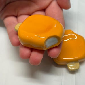 May include: Two orange, glossy, ceramic mushroom-shaped objects. Each has a small, light-colored stem and a rounded cap. One has a small chip on the side. The objects are held in a hand.