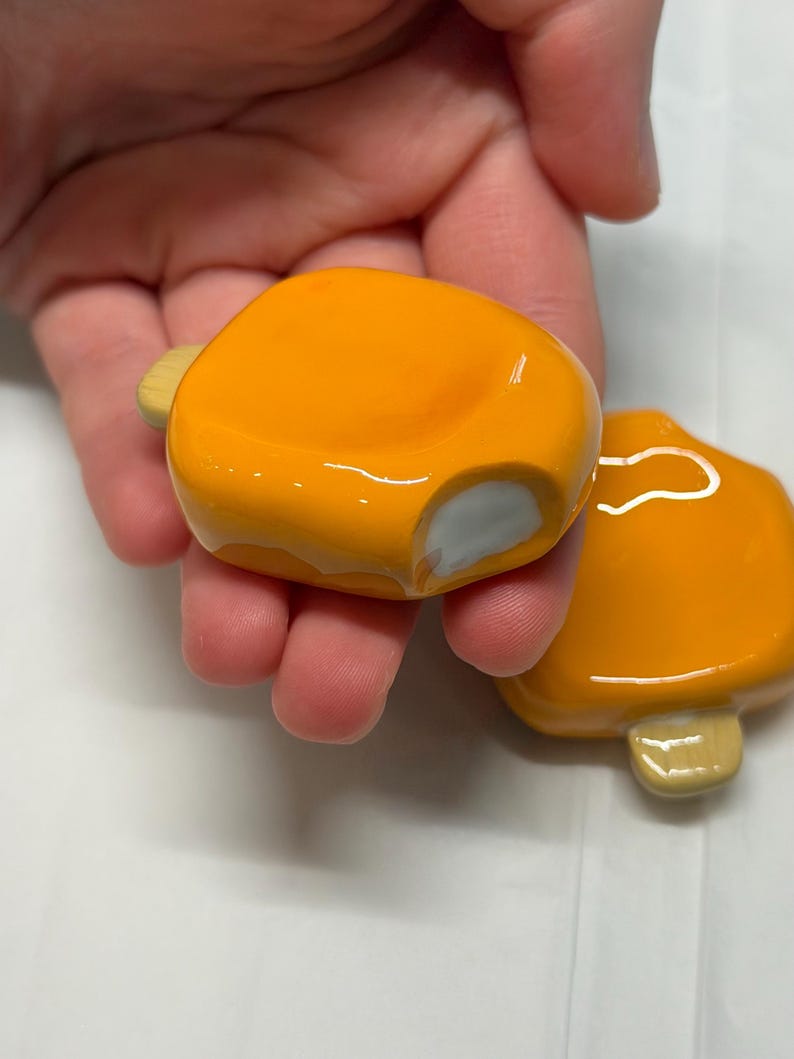 May include: Two orange, glossy, ceramic mushroom-shaped objects. Each has a small, light-colored stem and a rounded cap. One has a small chip on the side. The objects are held in a hand.