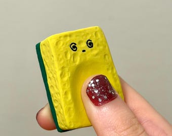 May include: A yellow sponge-shaped object with a cartoon face and a green edge. The object is held in a hand with a red nail with silver glitter.
