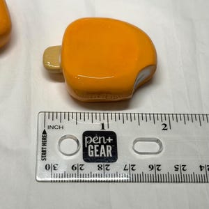 May include: An orange, popsicle-shaped glass bead with a light-colored stick. The bead is next to a ruler with the text "pen+ GEAR" and "INCH". The bead measures approximately 1 inch in length.