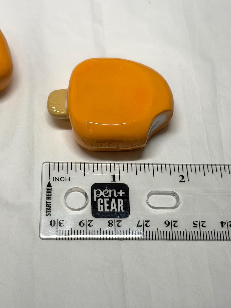 May include: An orange, popsicle-shaped glass bead with a light-colored stick. The bead is next to a ruler with the text "pen+ GEAR" and "INCH". The bead measures approximately 1 inch in length.