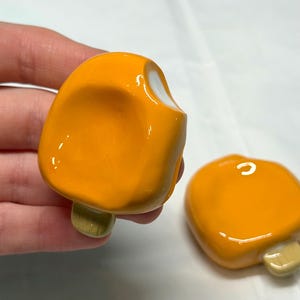 May include: Two orange popsicle-shaped ceramic pieces with a bite taken out of one. The popsicles have a glossy finish and a light green stick. One is held by a hand, the other is on a white surface.