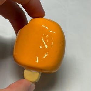 May include: A bright orange popsicle-shaped ceramic object with a light yellow base. The object is held by a hand against a white background. The popsicle has a glossy finish.