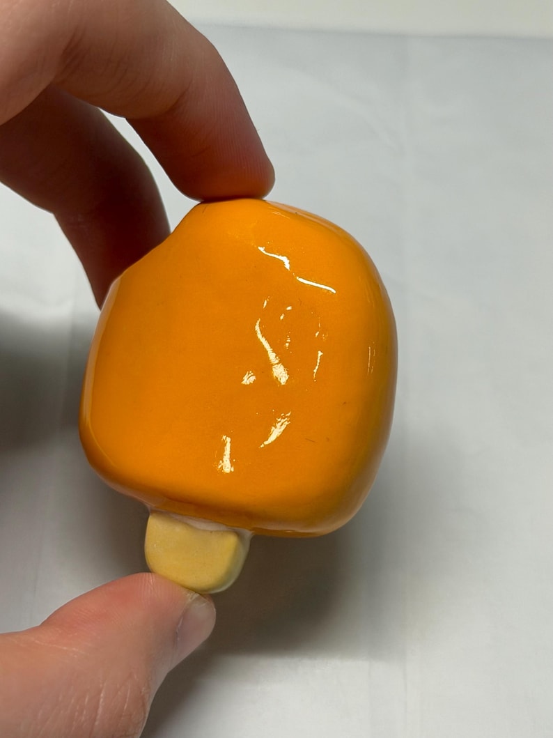 May include: A bright orange popsicle-shaped ceramic object with a light yellow base. The object is held by a hand against a white background. The popsicle has a glossy finish.