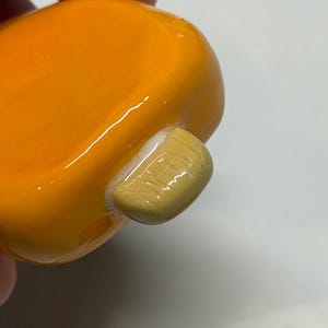May include: Close-up of a small, rounded, orange object with a light tan, rectangular attachment. The object has a glossy finish and appears to be made of ceramic or a similar material. The attachment has a textured surface.