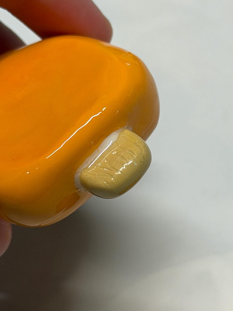 May include: Close-up of a small, rounded, orange object with a light tan, rectangular attachment. The object has a glossy finish and appears to be made of ceramic or a similar material. The attachment has a textured surface.