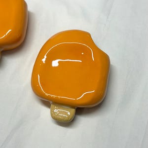 May include: Close-up of two orange popsicle-shaped objects with a glossy finish. Each has a light brown stick. The objects are on a white surface. The popsicles are a bright, solid orange color.