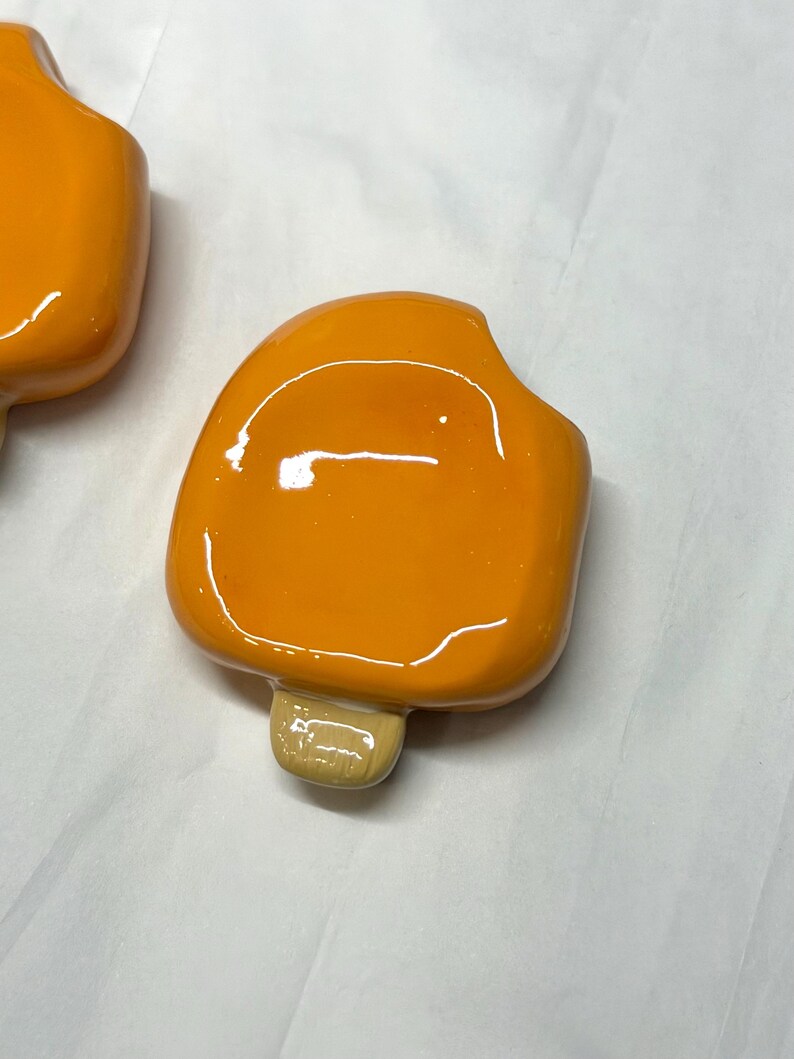 May include: Close-up of two orange popsicle-shaped objects with a glossy finish. Each has a light brown stick. The objects are on a white surface. The popsicles are a bright, solid orange color.