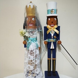 Puede incluir: Un par de figuras de cascanueces de madera vestidas de novia y novio. La novia lleva un vestido blanco de encaje con una faja azul y una corona dorada. El novio lleva un traje azul con ribetes dorados y un sombrero de copa blanco.