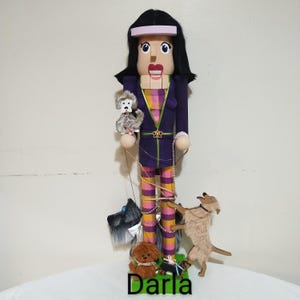 May include: A wooden nutcracker doll dressed in a purple and pink striped outfit with a pink headband. The doll is holding a leash and walking four small dogs. The dogs are a white poodle, a black terrier, a brown poodle, and a tan terrier. The doll is labeled "Darla".
