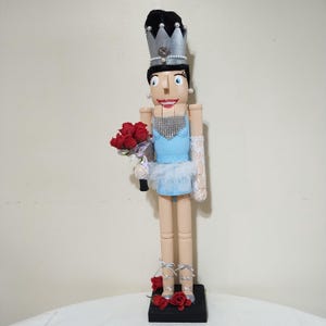 May include: A wooden nutcracker figurine of a ballerina wearing a light blue dress, a silver crown, and holding a bouquet of red roses. The ballerina has a white tulle skirt and silver shoes.