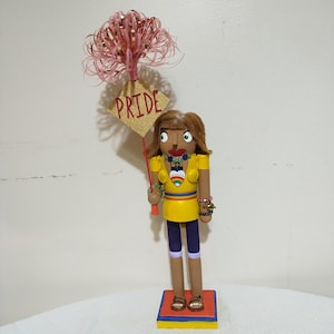 Puede incluir: Una muñeca cascanueces de madera con piel morena, que lleva un vestido amarillo con rayas arcoíris, un corazón arcoíris y un collar de cuentas verdes. La muñeca sostiene un cartel que dice "PRIDE" y tiene un boa de plumas rosa en la cabeza.