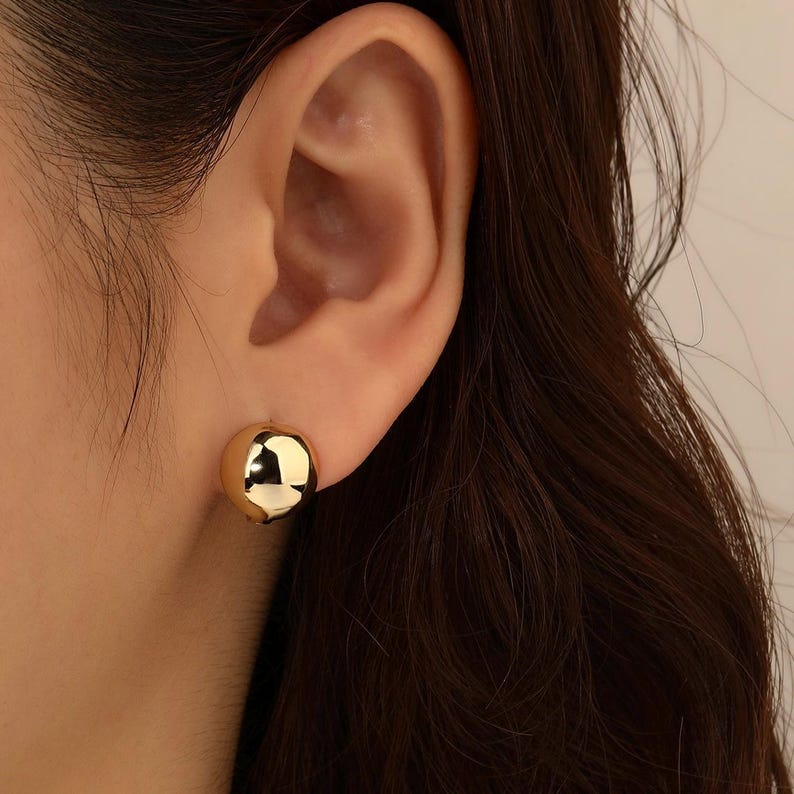 May include: A close-up of a gold-coloured, spherical stud earring. The earring is smooth and reflective, catching the light. The earring is worn on an ear, with dark hair visible in the background. The earring is a simple, yet elegant piece of jewellery.