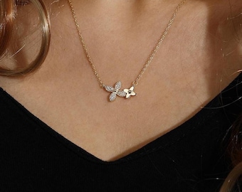 May include: A delicate gold necklace featuring two flower pendants. One pendant is adorned with sparkling clear crystals, while the other is a smaller, solid gold flower. The necklace is displayed on a black top.