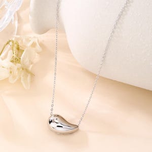 May include: A silver-colored necklace with a teardrop-shaped pendant. The pendant is smooth and reflective, hanging from a delicate chain. The necklace is displayed against a soft, neutral background, highlighting its minimalist design.
